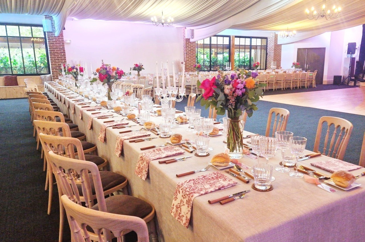 Grande table de réception décorée avec des fleurs colorées, des chandeliers et des couverts alignés dans une salle lumineuse.