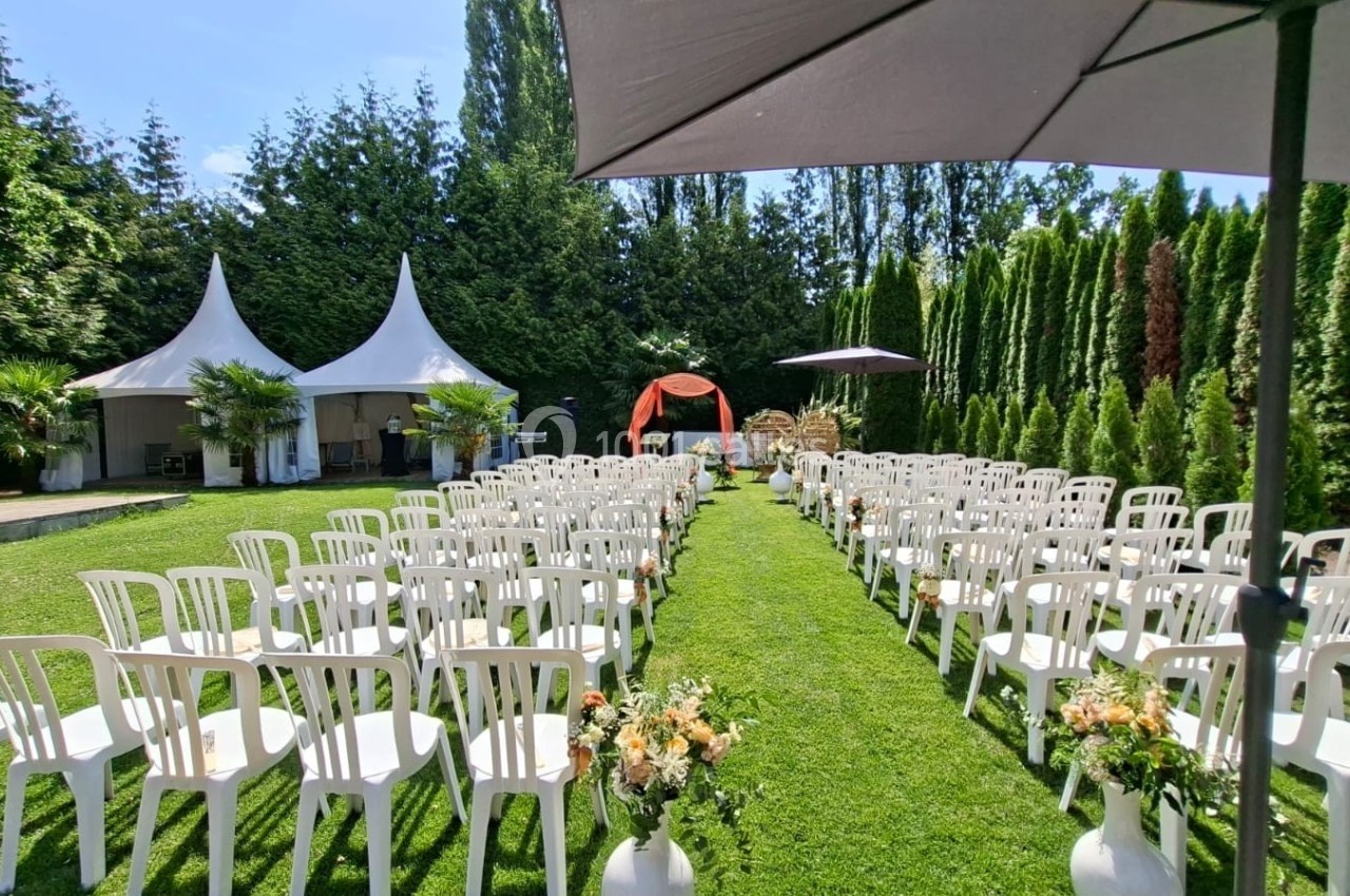 Chaises blanches alignées en extérieur pour une cérémonie, avec une arche décorée et des tentes en arrière-plan.