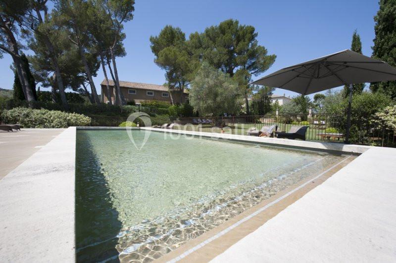 Piscine extérieure entourée de végétation, avec parasol, transats et maisons en arrière-plan sous un ciel dégagé.