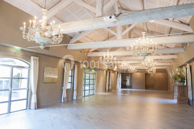 Salle lumineuse avec poutres apparentes, lustres suspendus, grandes fenêtres et sol en carrelage clair.