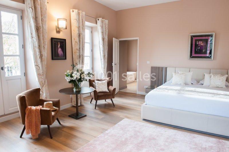 Chambre lumineuse avec lit double, fauteuils, table ronde, bouquet de fleurs, grandes fenêtres et décor élégant.