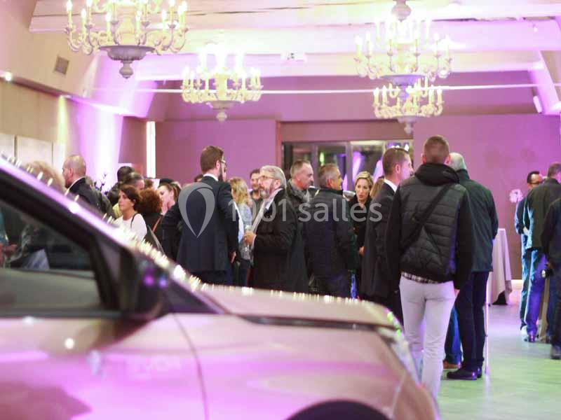 Groupe de personnes discutant dans une salle éclairée par des lustres, avec une voiture visible au premier plan.