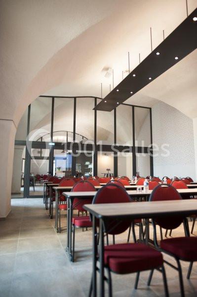 Salle lumineuse avec des tables et des chaises rouges alignées, sous un plafond voûté et éclairée par des suspensions…