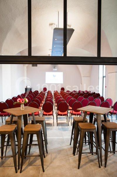 Salle de conférence avec rangées de chaises rouges alignées, écran de projection et tables hautes au premier plan.