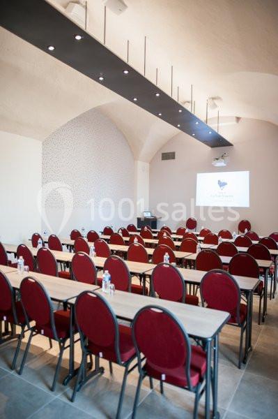 Salle de conférence lumineuse avec des rangées de tables en bois et de chaises rouges, écran de projection allumé au fond.