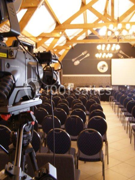 Salle de conférence vide avec des rangées de chaises, une caméra de tournage et un écran de projection en arrière-plan.