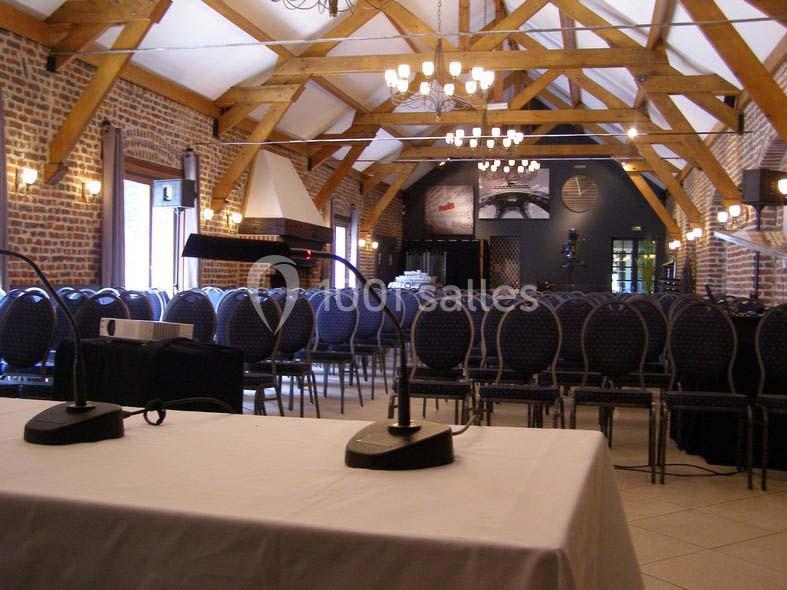 Salle de conférence avec des rangées de chaises, une table équipée de micros et un plafond en poutres apparentes.