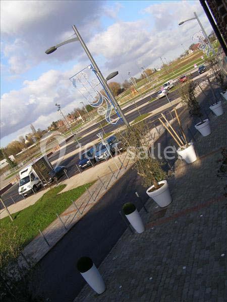 Vue d'un parking vide bordé de lampadaires et de pots de fleurs blancs, avec un ciel partiellement nuageux.