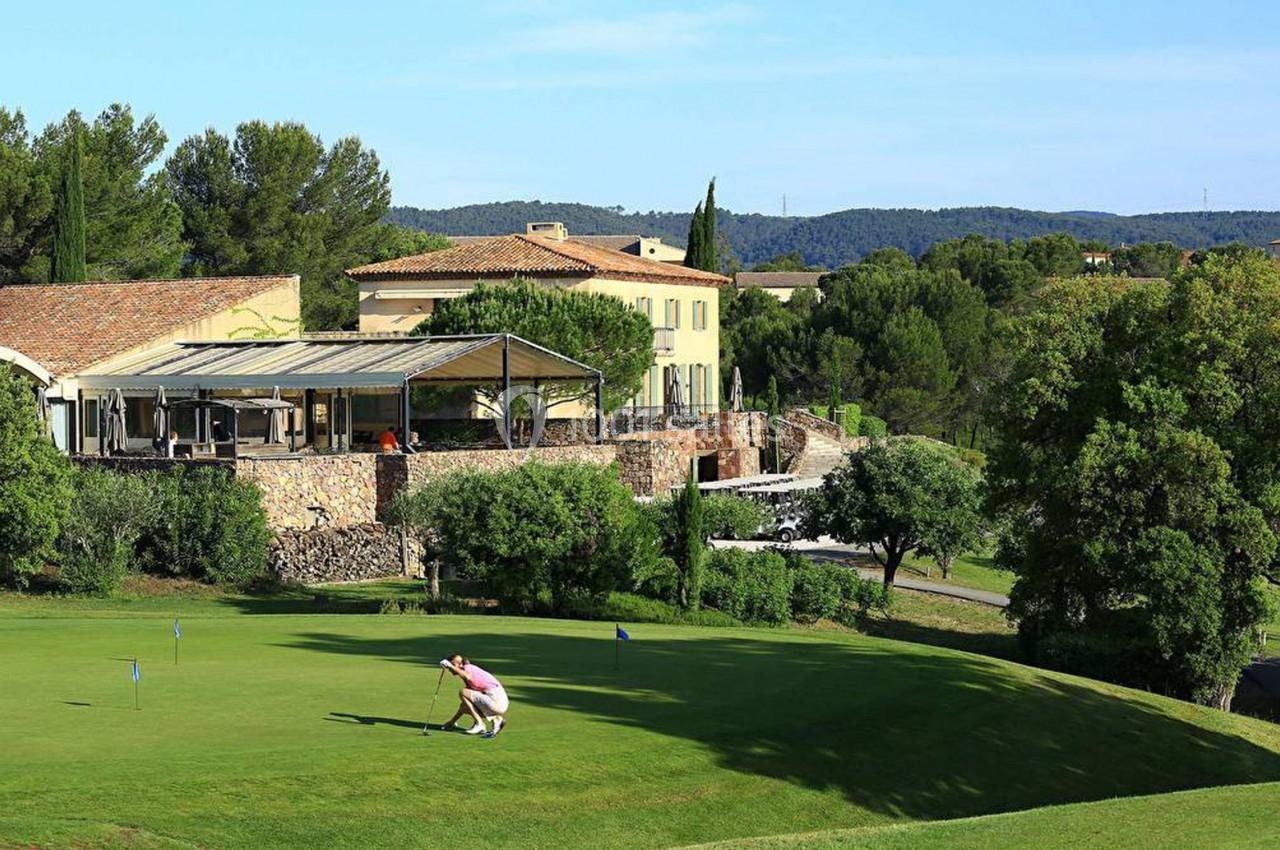 Un golfeur ajuste son coup sur un green entouré de végétation, avec des bâtiments en arrière-plan.