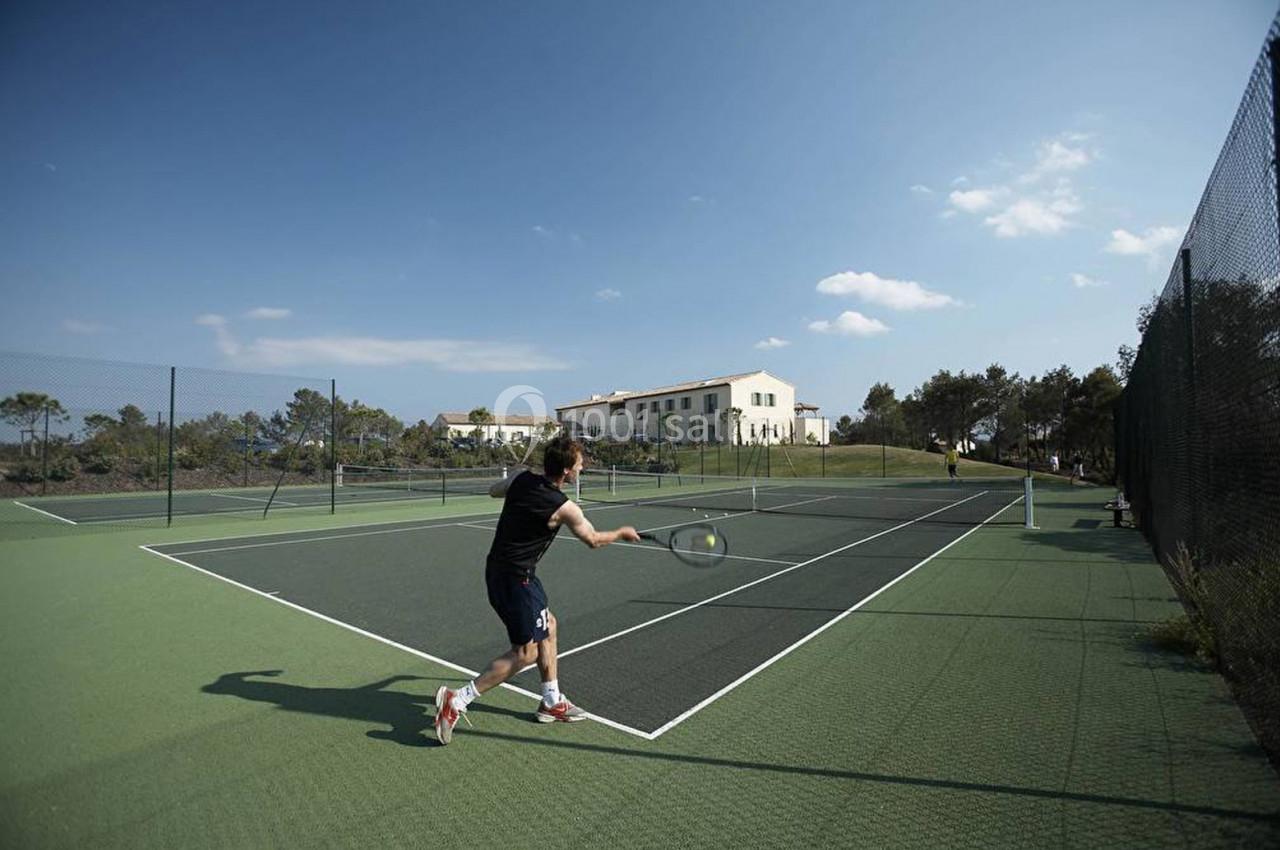 Un homme joue au tennis sur un court extérieur entouré de clôtures, avec un bâtiment et des arbres en arrière-plan.