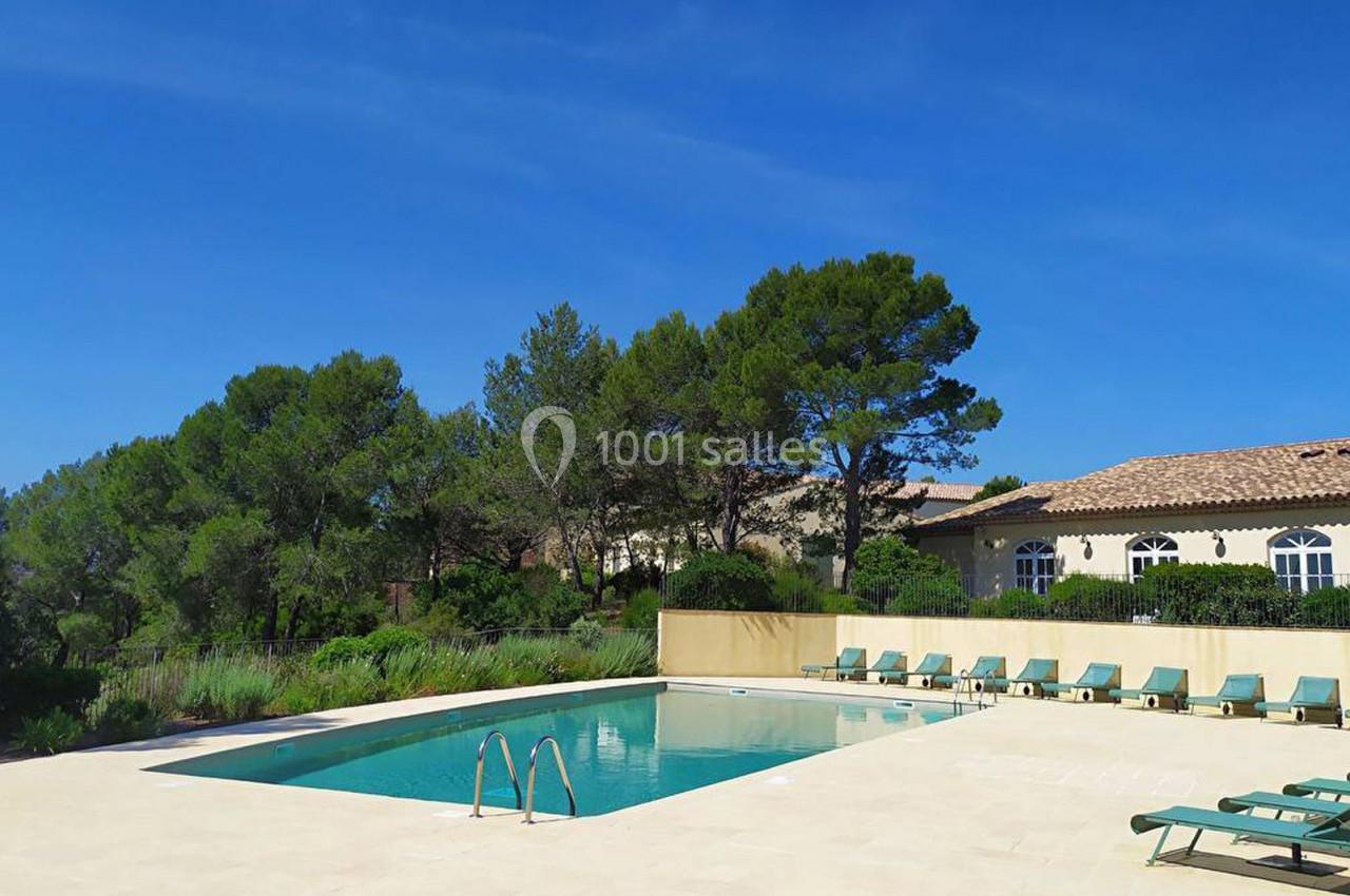 Piscine extérieure entourée de chaises longues, bordée de végétation et d'un bâtiment avec toit en tuiles sous un ciel bleu.