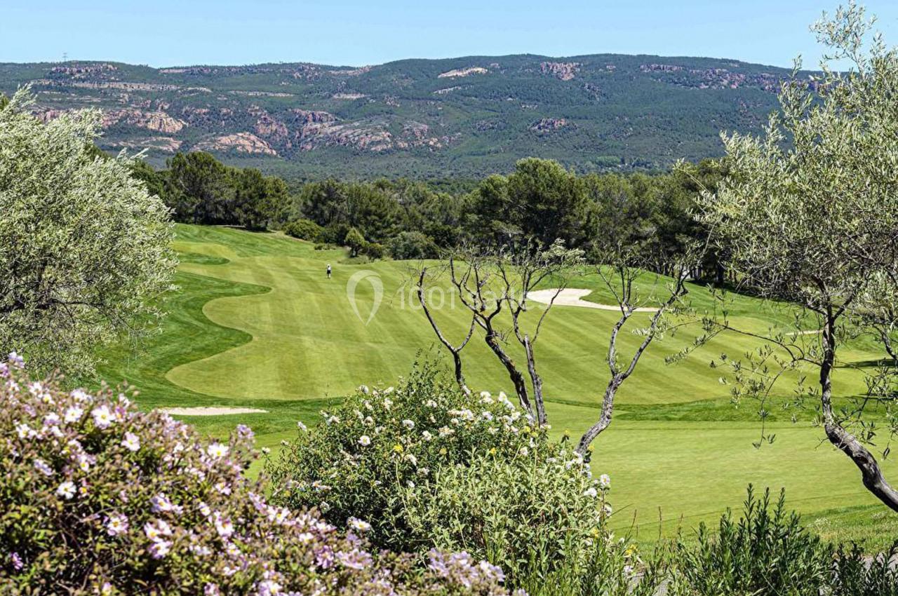 Paysage de golf avec pelouse verdoyante, arbres, buissons fleuris et collines en arrière-plan sous un ciel clair.