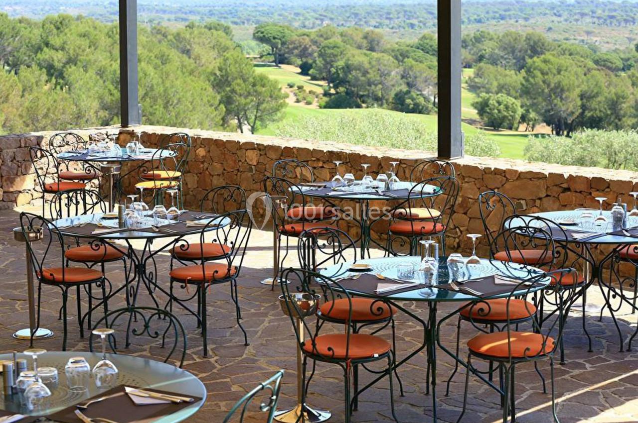 Terrasse d'un restaurant avec des tables en fer forgé, vue sur un paysage arboré et un terrain de golf.