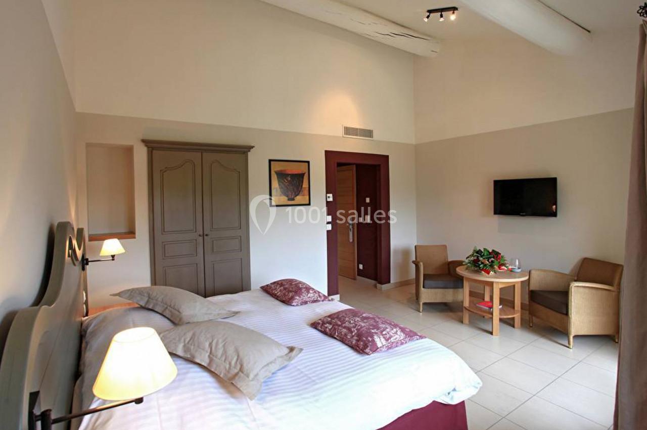 Chambre lumineuse avec lit double, armoire, télévision murale, table ronde et deux fauteuils dans un décor sobre.