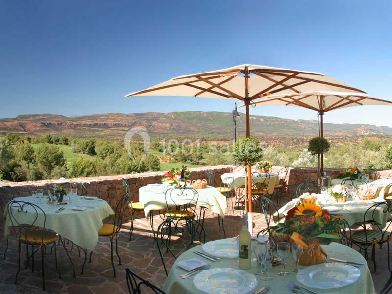 Terrasse ensoleillée avec tables dressées, parasols et vue sur un paysage vallonné verdoyant et aride.
