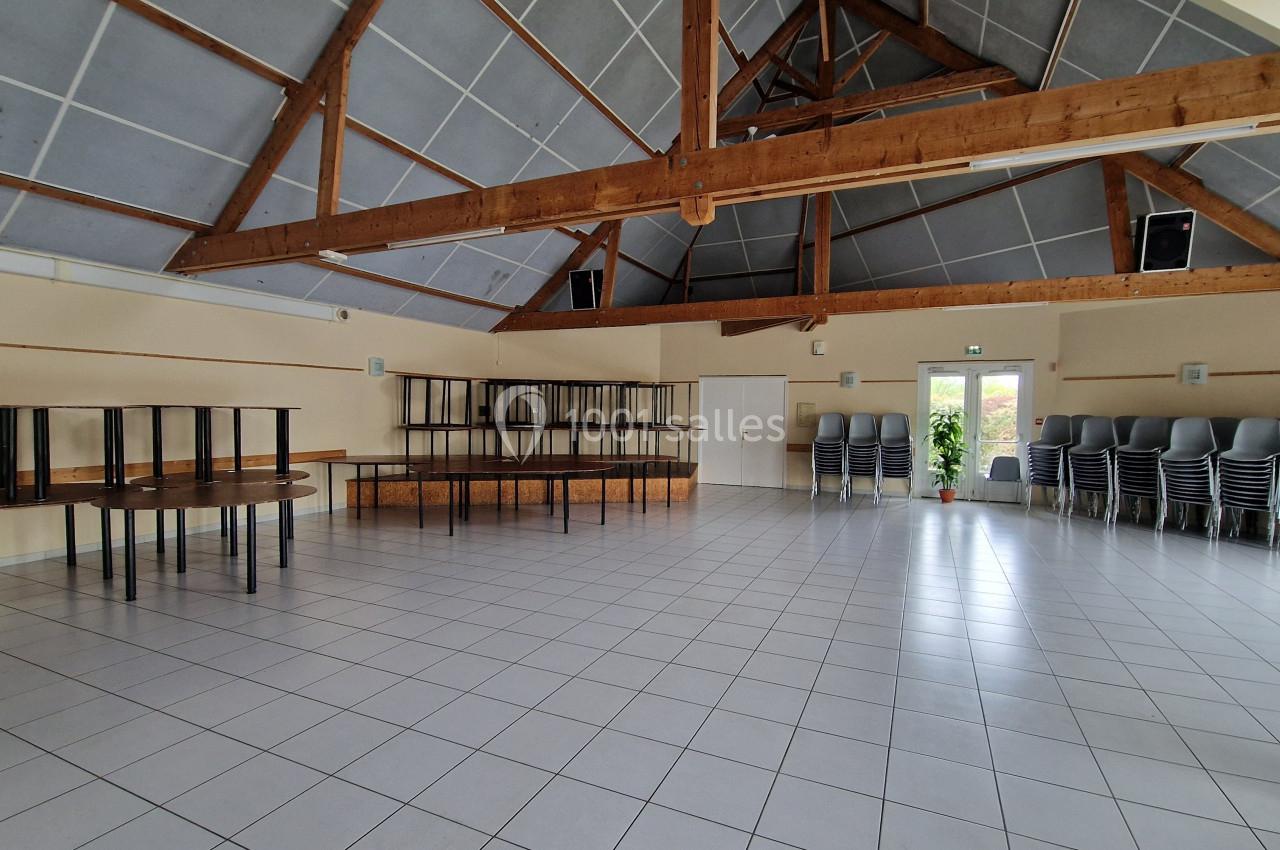 Salle vide avec un sol carrelé, des poutres apparentes, des tables empilées et des chaises rangées contre un mur.