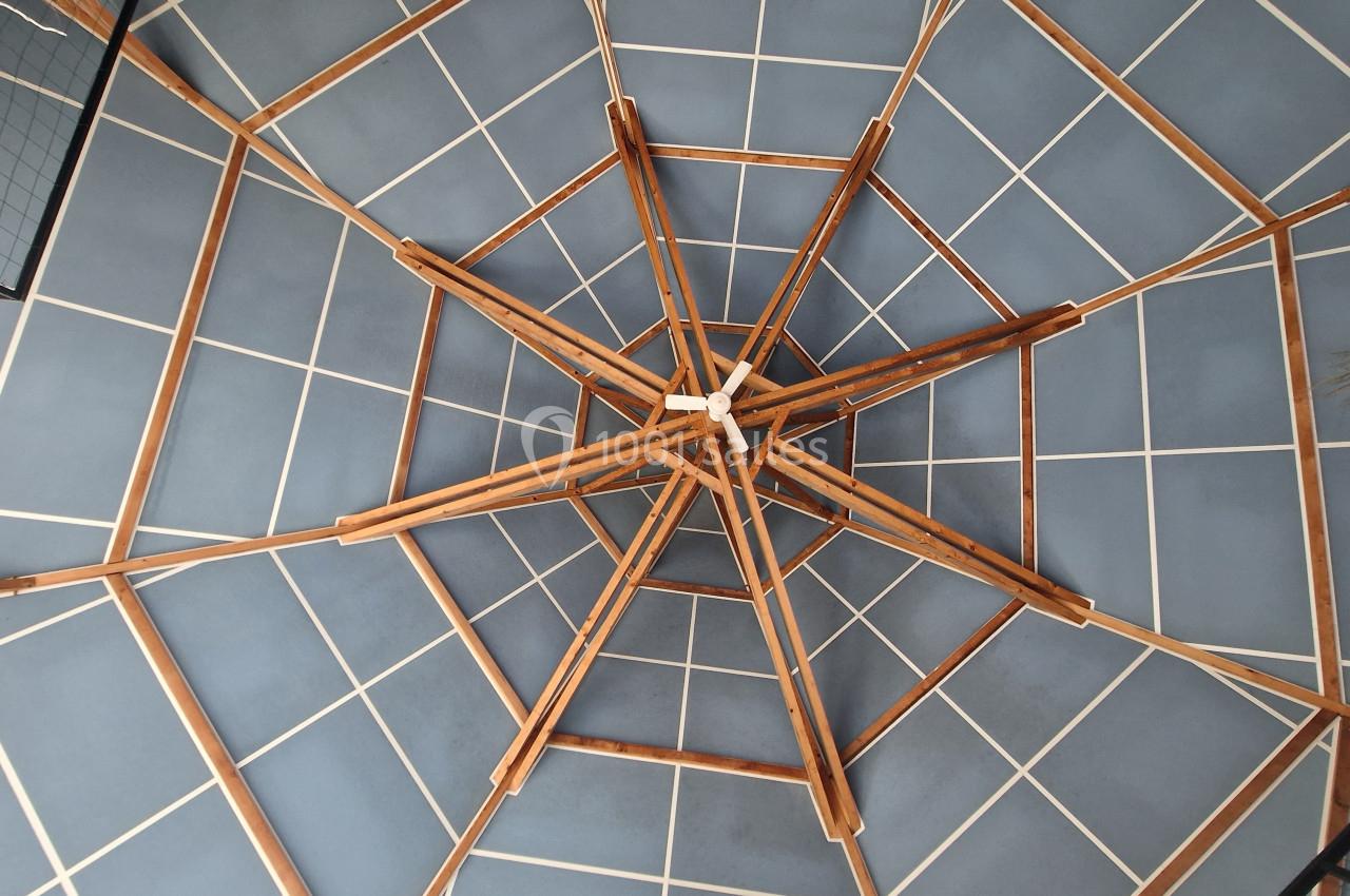 Structure en bois formant un motif géométrique complexe vue depuis le dessous, avec un plafond bleu quadrillé.