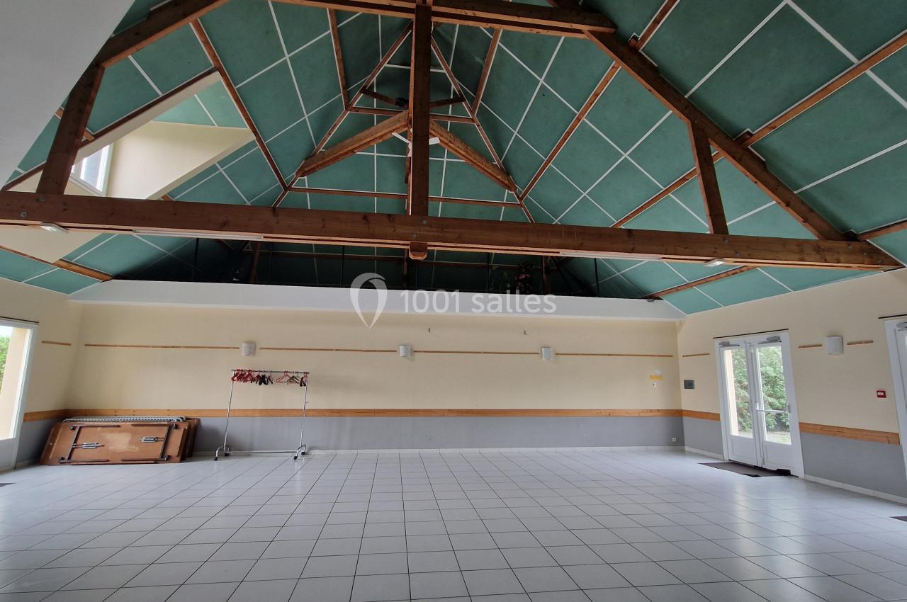Salle vide avec plafond en charpente apparente, murs clairs, carrelage blanc et grandes fenêtres laissant entrer la lumière.