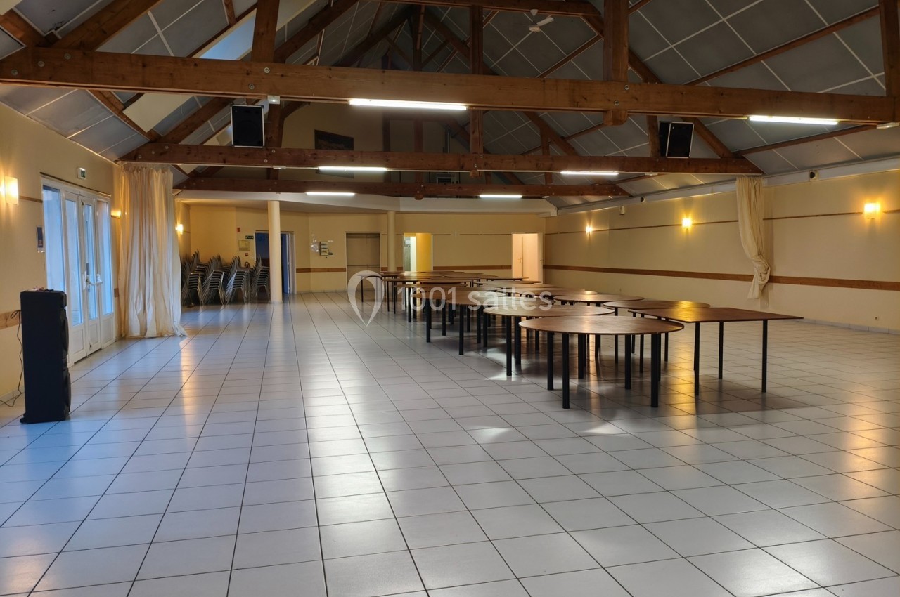 Salle spacieuse avec charpente en bois apparente, tables alignées et chaises empilées dans un coin.