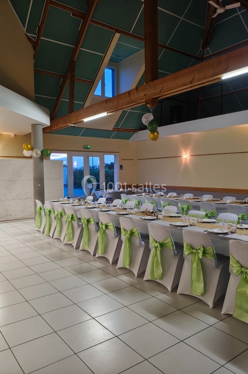 Salle décorée pour un événement, avec des tables alignées, nappes blanches et chaises ornées de nœuds verts.