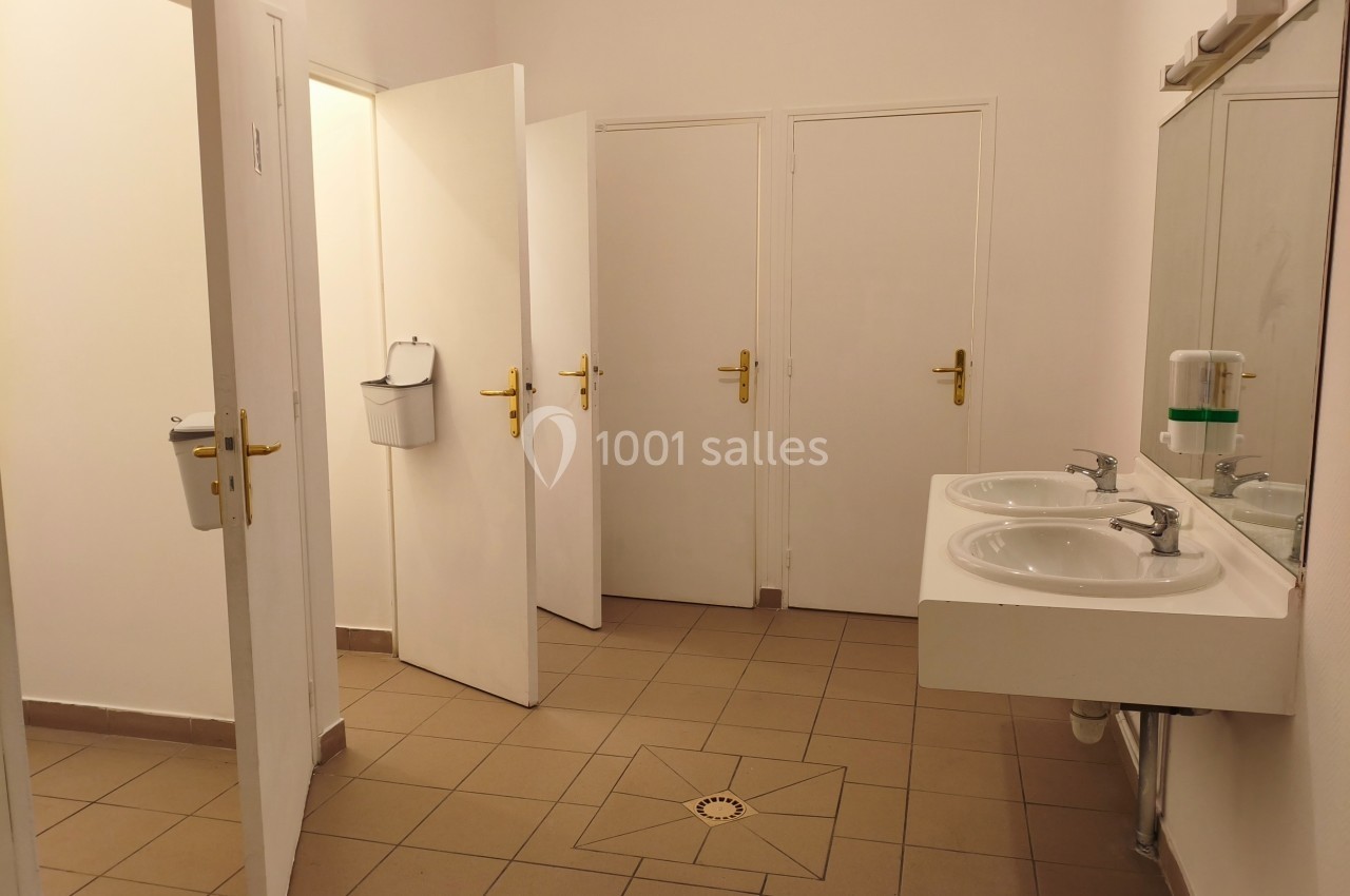 Salle de bains avec trois portes, deux lavabos blancs, un miroir et un distributeur de savon sur un carrelage beige.