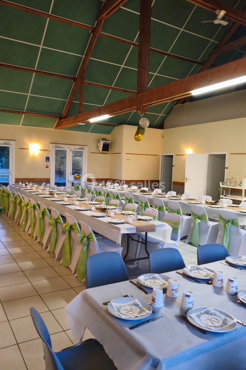 Salle de réception décorée avec des tables alignées, nappes blanches, chaises ornées de rubans verts.