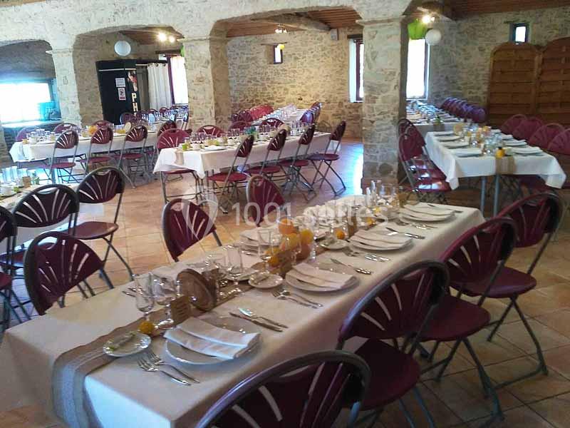 Salle en pierre aménagée avec des tables dressées pour un repas, chaises rouges pliantes et décoration sobre.