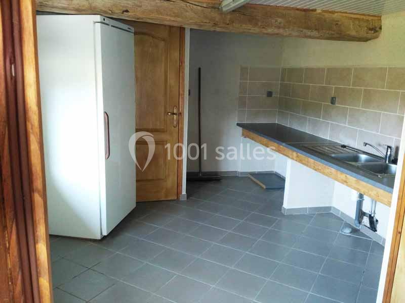 Cuisine vide avec carrelage gris, plan de travail en bois, évier inox, réfrigérateur blanc et poutre apparente au plafond.