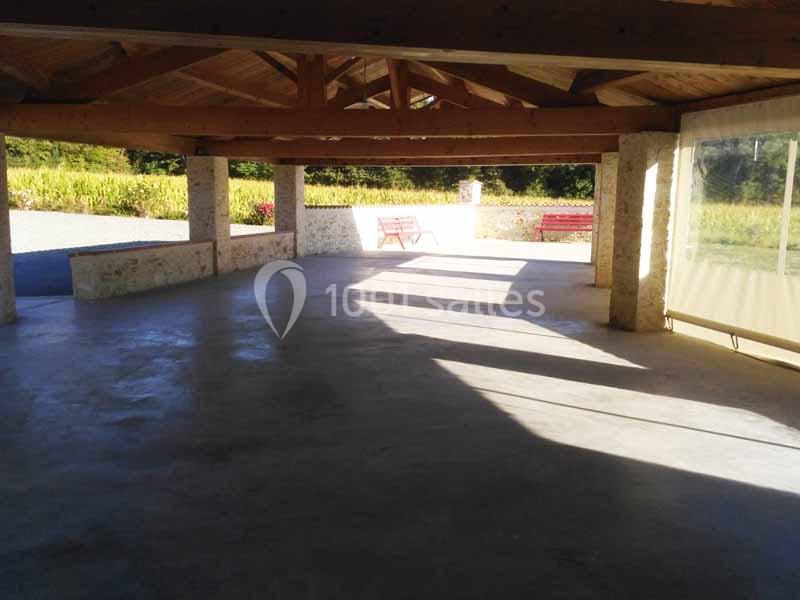 Abri ouvert avec charpente en bois, sol en béton et vue sur un espace extérieur avec bancs et végétation.