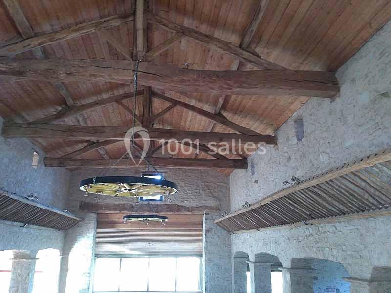 Plafond en bois avec poutres apparentes dans une salle en pierre, éclairée par des fenêtres et des lustres suspendus.