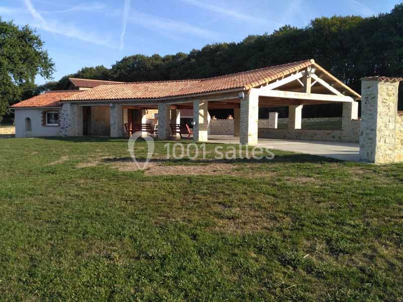 Bâtiment en pierre avec toit en tuiles et espace couvert, entouré de pelouse et d'arbres en arrière-plan.