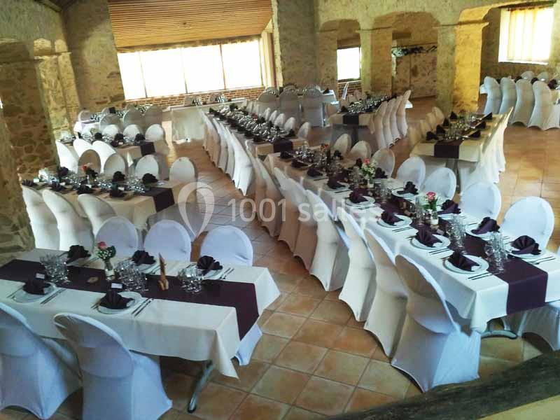 Salle de réception avec tables alignées, nappes blanches, chemins de table sombres et chaises couvertes.