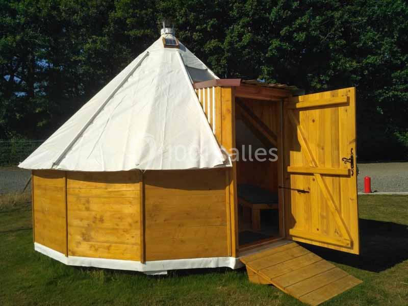 Yourte en bois avec une porte ouverte et une rampe, située sur une pelouse devant des arbres.