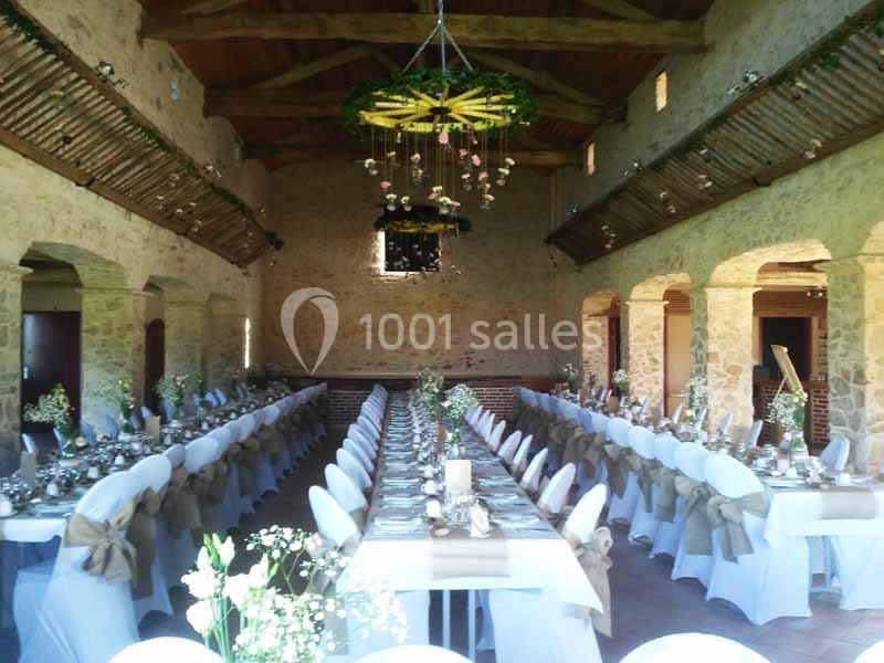 Salle de réception en pierre avec poutres apparentes, tables longues décorées de nappes blanches et fleurs.