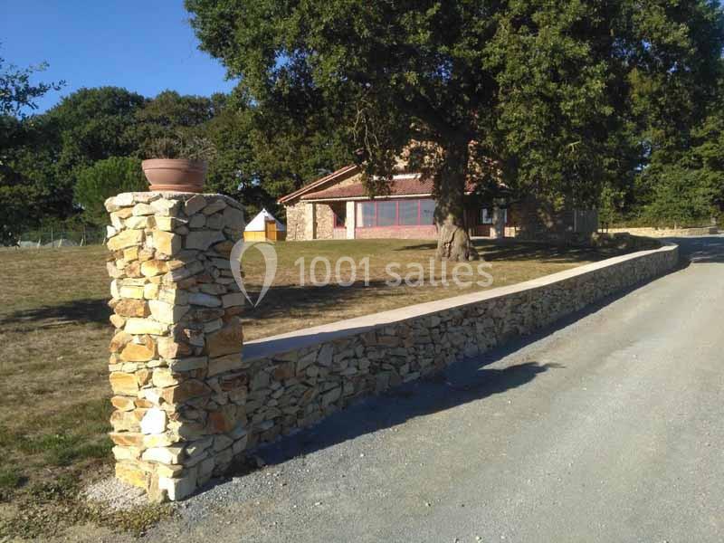 Mur en pierre sèche longeant une allée, avec une maison en pierre et un grand arbre en arrière-plan.
