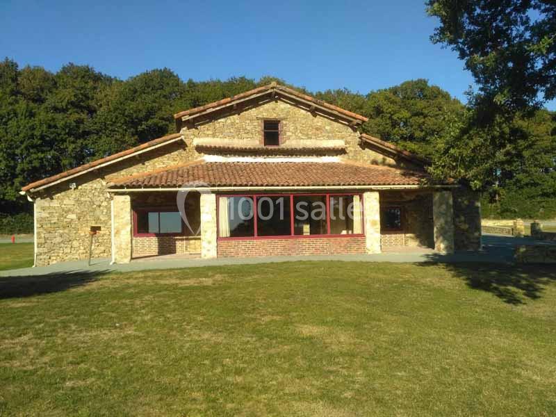 Maison en pierre avec toit en tuiles, grandes baies vitrées et pelouse à l'avant, entourée d'arbres.