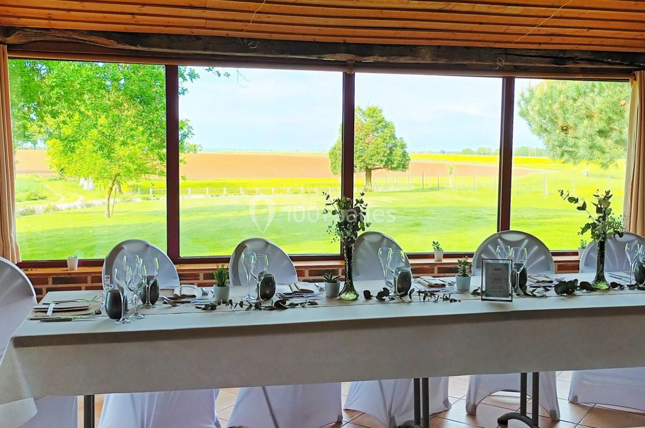 Table dressée avec nappes blanches, décorations végétales et vue sur un paysage champêtre à travers de grandes baies vitrées.