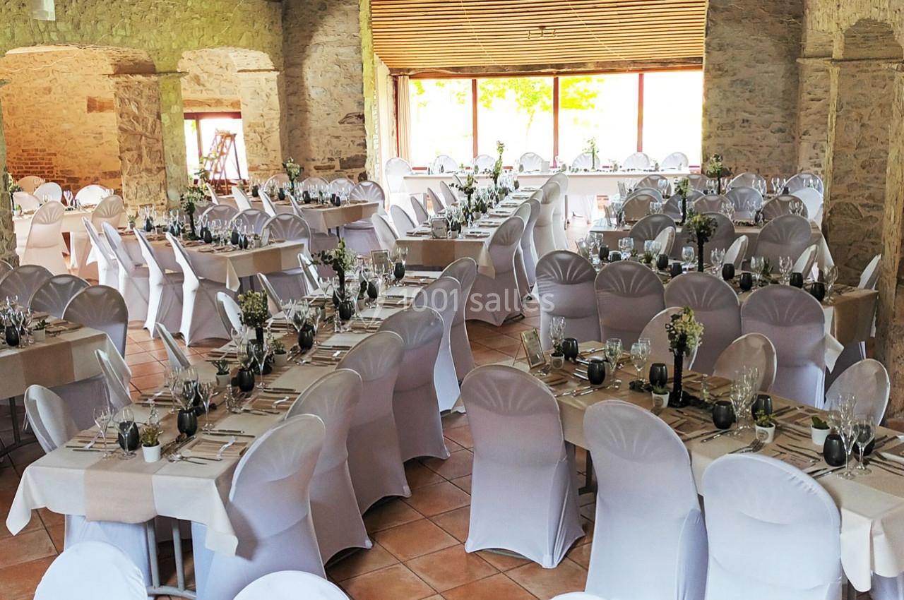 Salle de réception aménagée avec des tables dressées et des chaises blanches, dans un cadre en pierre avec lumière naturelle.