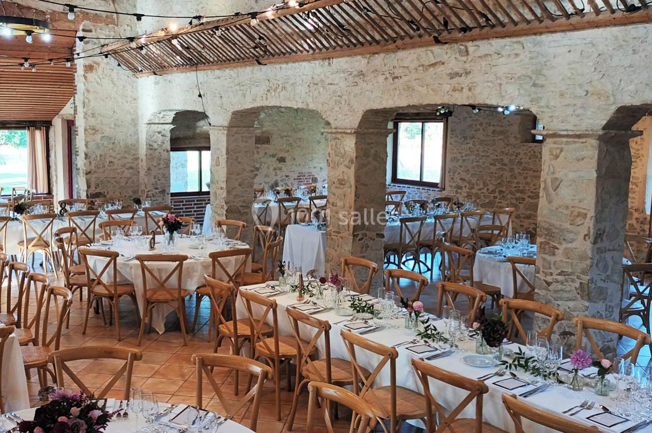 Salle en pierre aménagée pour un événement, avec des tables décorées, des chaises en bois et des guirlandes lumineuses.