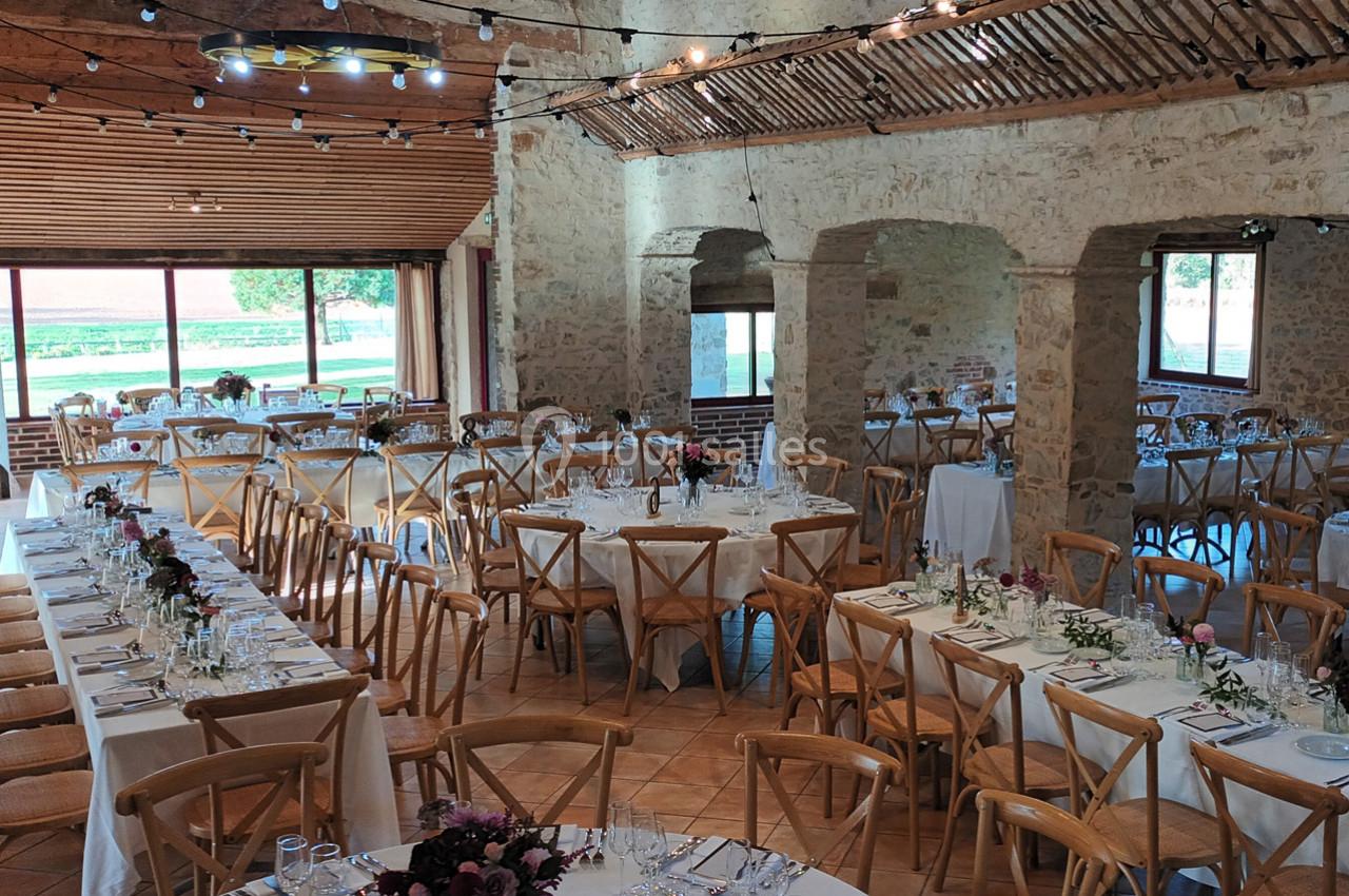 Salle de réception lumineuse avec tables décorées pour un événement, entourée de murs en pierre et grandes fenêtres donnant…