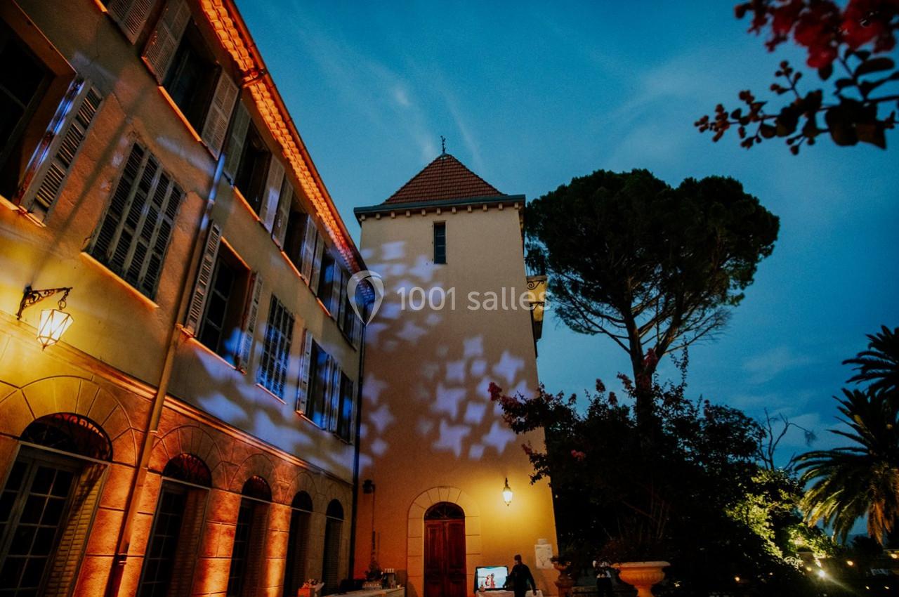 Façade d'un bâtiment éclairé par des lumières colorées au crépuscule, entouré d'arbres et de végétation.