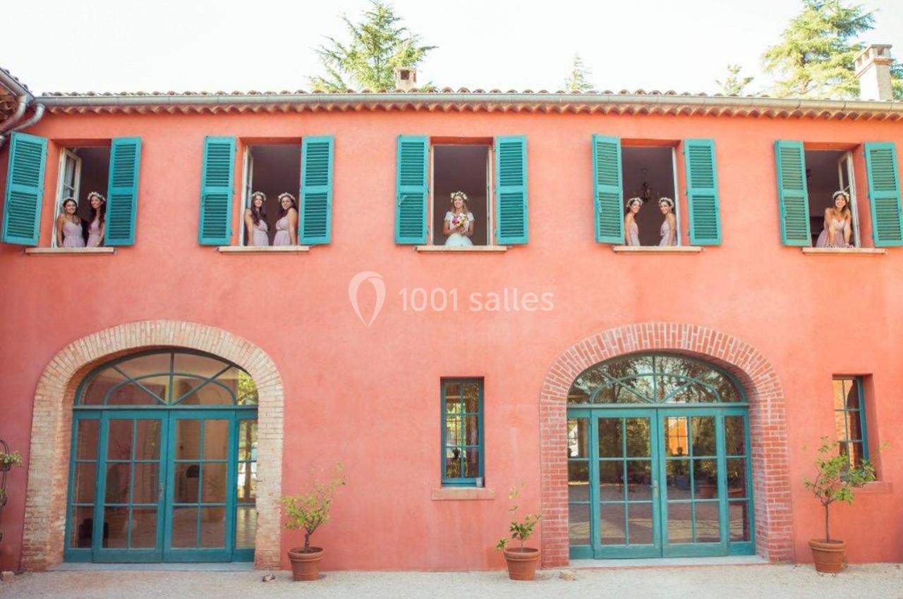 Façade d'une maison rose avec des volets verts, des personnes regardant par les fenêtres du premier étage.