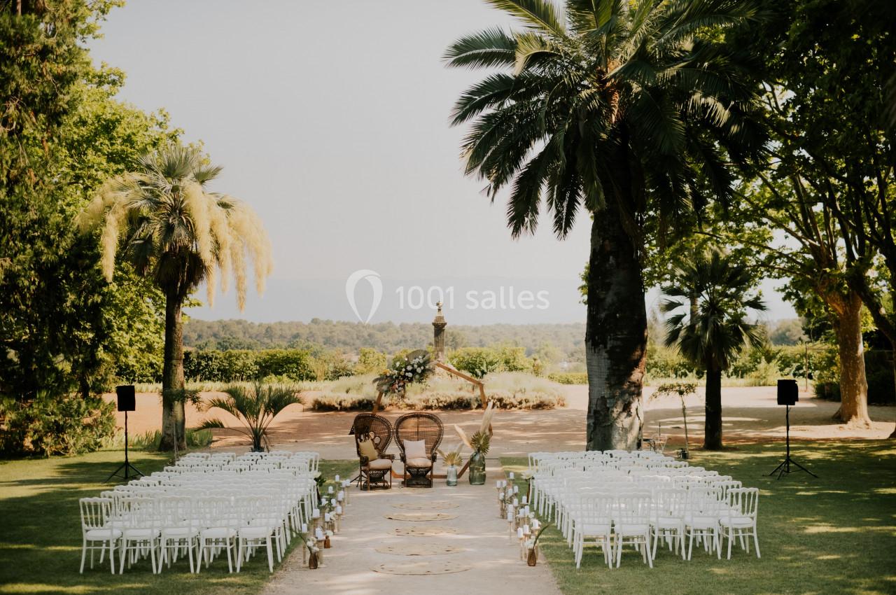 Allée centrale bordée de chaises blanches menant à deux fauteuils en osier, dans un jardin avec palmiers et fontaine.