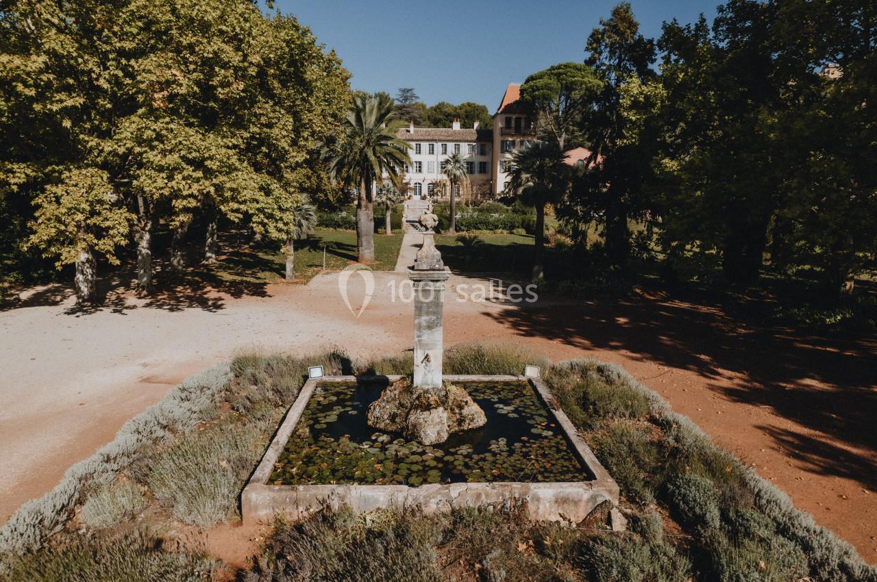 Fontaine entourée de végétation dans un jardin, avec un bâtiment ancien en arrière-plan sous un ciel dégagé.