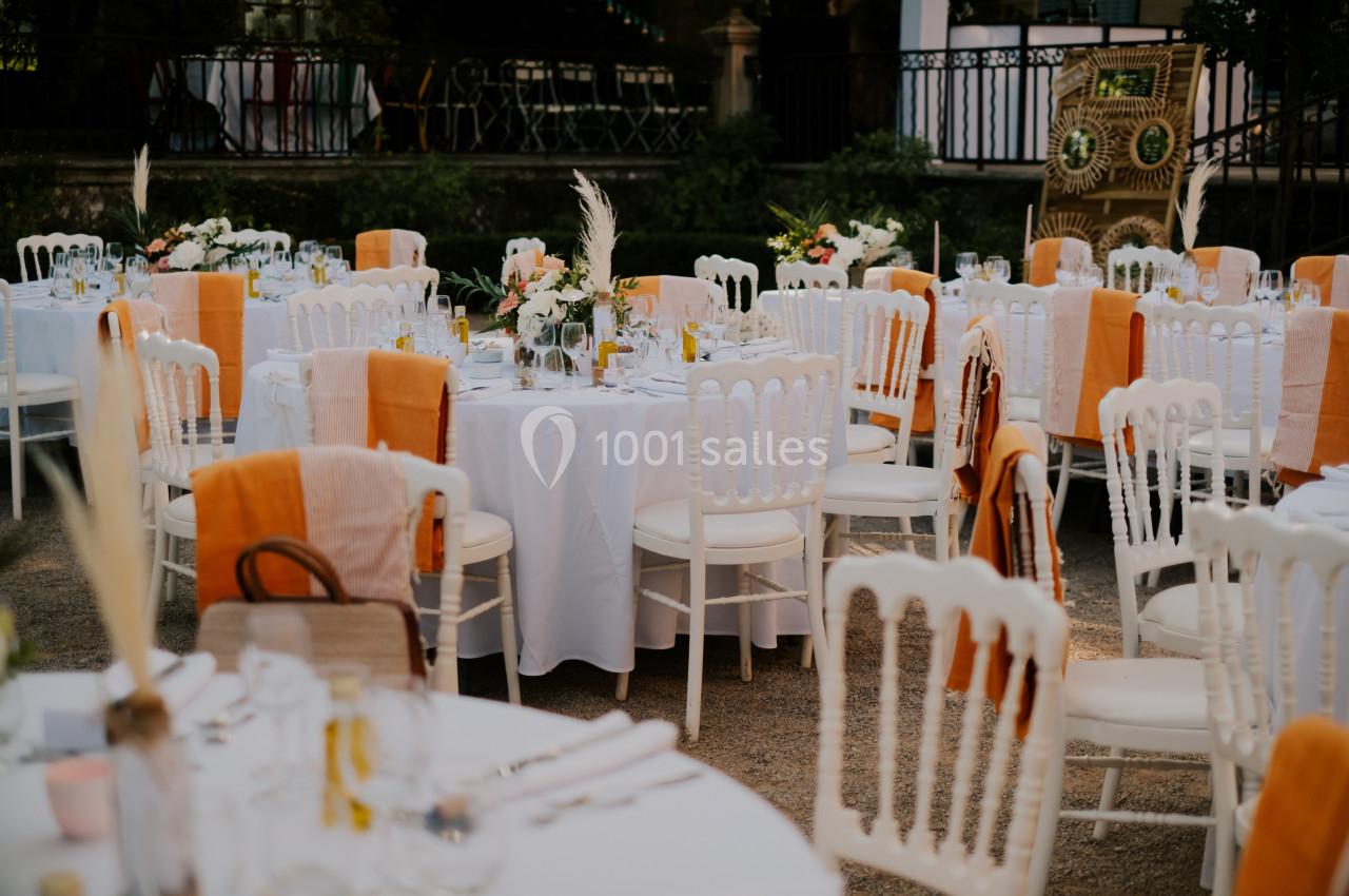Tables rondes décorées de nappes blanches et chaises blanches avec des touches orange, disposées en extérieur.