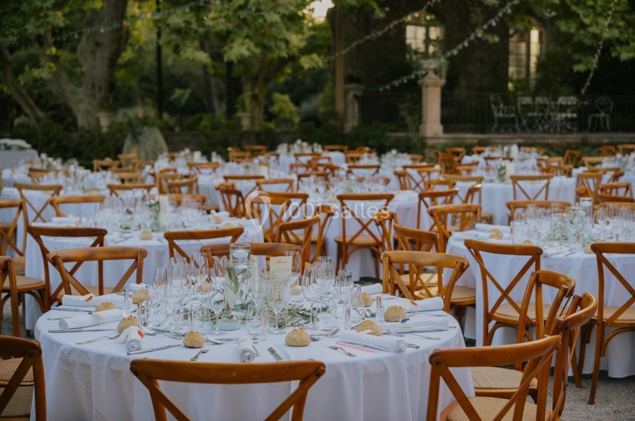 Tables rondes dressées avec nappes blanches, chaises en bois, vaisselle et décoration florale dans un jardin extérieur.