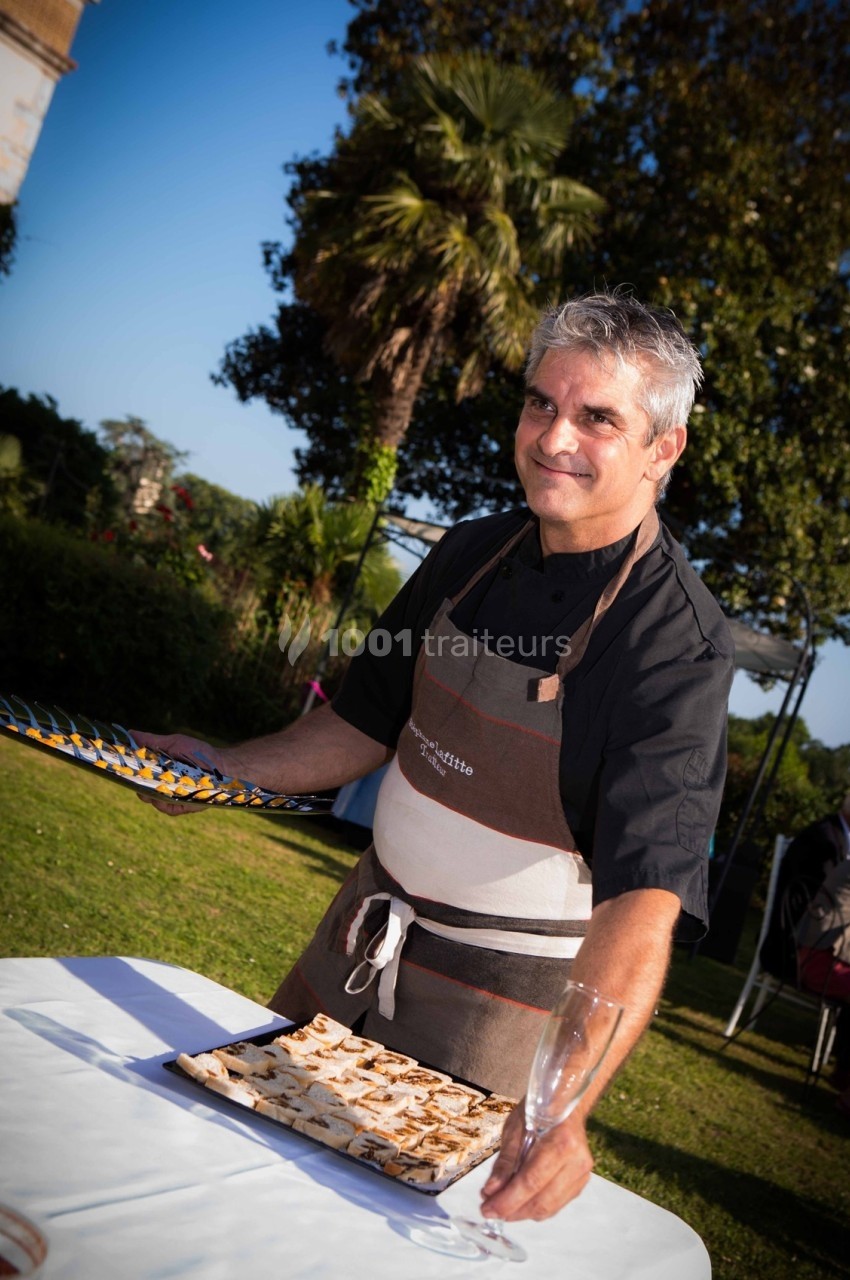 Un homme en tablier sert un plateau de petits fours sur une table en extérieur, avec un jardin en arrière-plan.