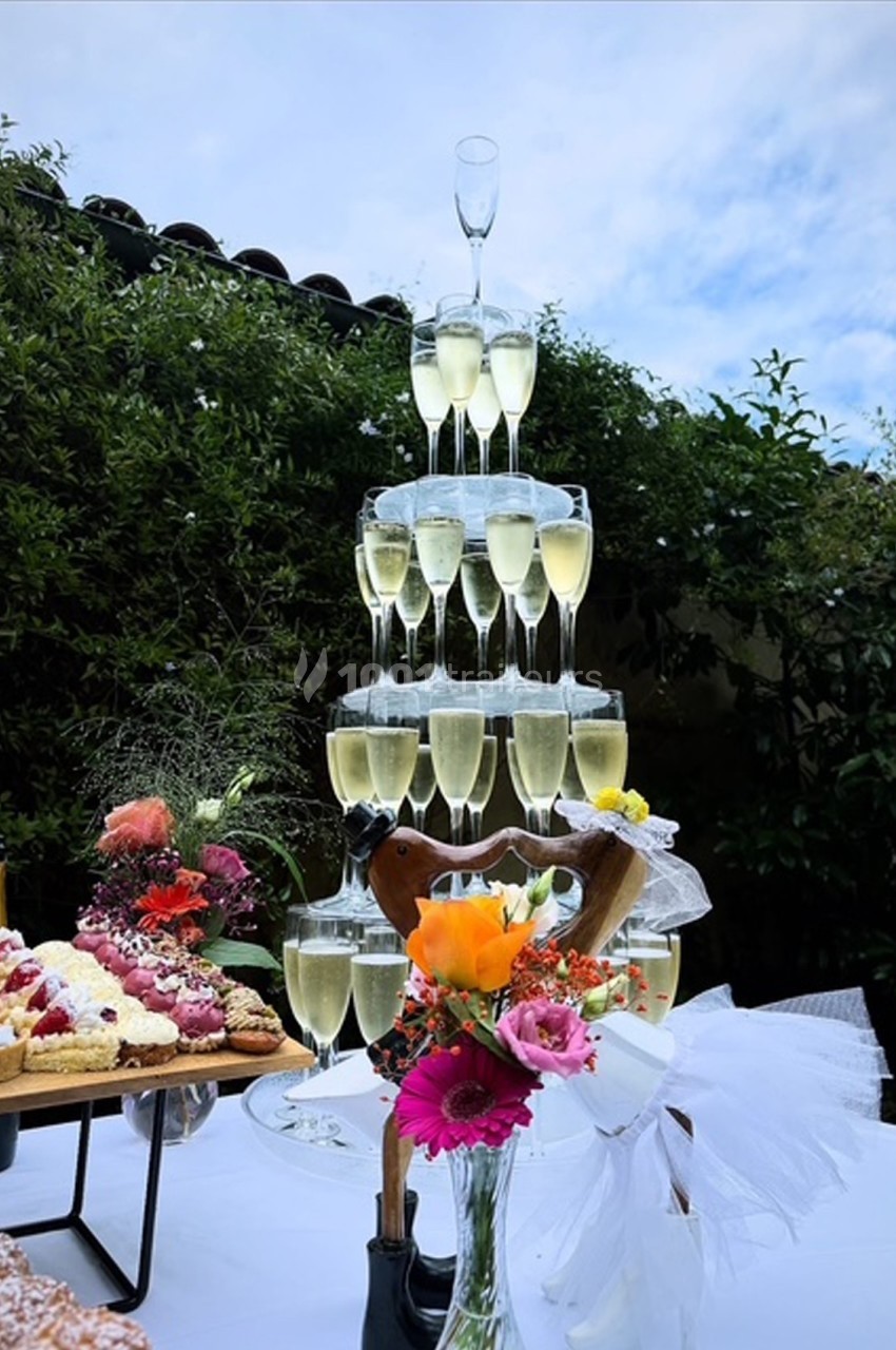 Tour de coupes de champagne empilées en pyramide sur une table décorée de fleurs et de desserts, en extérieur.