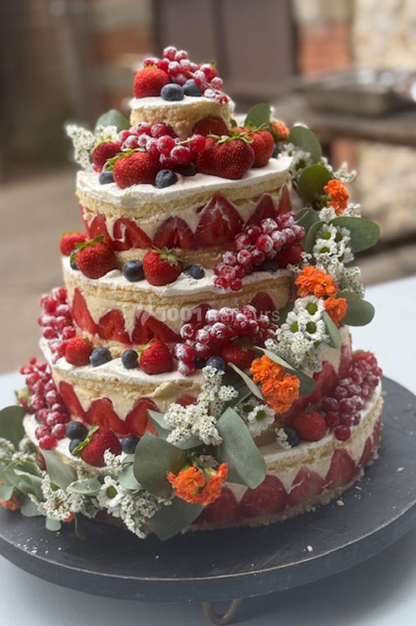 Gâteau de mariage à étages décoré de fruits rouges, baies, fleurs fraîches et feuillage, présenté sur un plateau noir.
