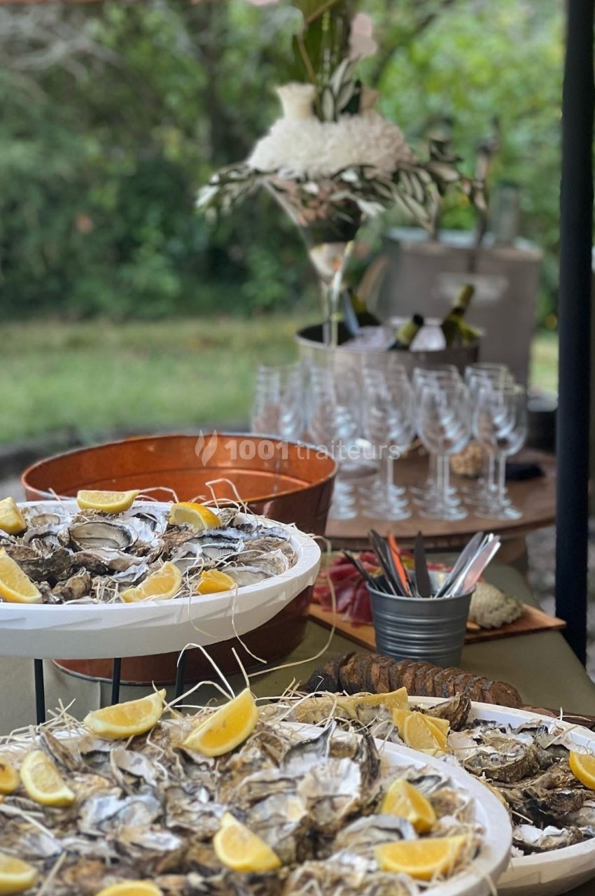 Plateaux d'huîtres garnis de quartiers de citron sur une table extérieure, avec verres et bouteilles en arrière-plan.